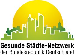 Abgebildet ist das Logo des Gesunde Städte-Netzwerks. Dieses ist die grüne Skyline einer Stadt vor einer großen gelben Sonne am Horizont. Logo des Gesunde Städte-Netzwerk