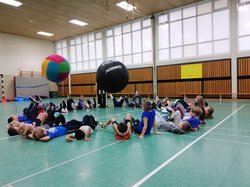 Zu sehen sind Kinder, die in einer Turnhalle im Kreis auf Rücken liegen und zwei große Bälle mit den Füßen hin- und her treten. Foto: SSB e.V.