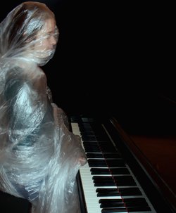 Eine Frau unter einer Plastikfolie spielt Klavier. Foto: Gila Schmidt/Kiyan Naderi