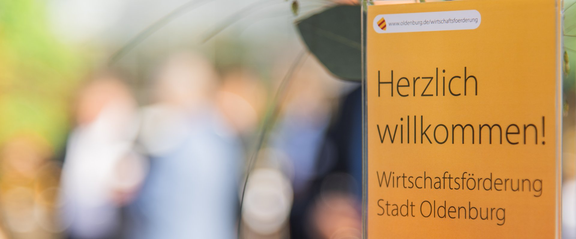 Im Vordergrund: Tischschild „Herzlich willkommen, Wirtschaftsförderung Stadt Oldenburg“, im Hintergrund: Gäste einer Veranstaltung der Wirtschaftsförderung. Foto: Imke Folkerts