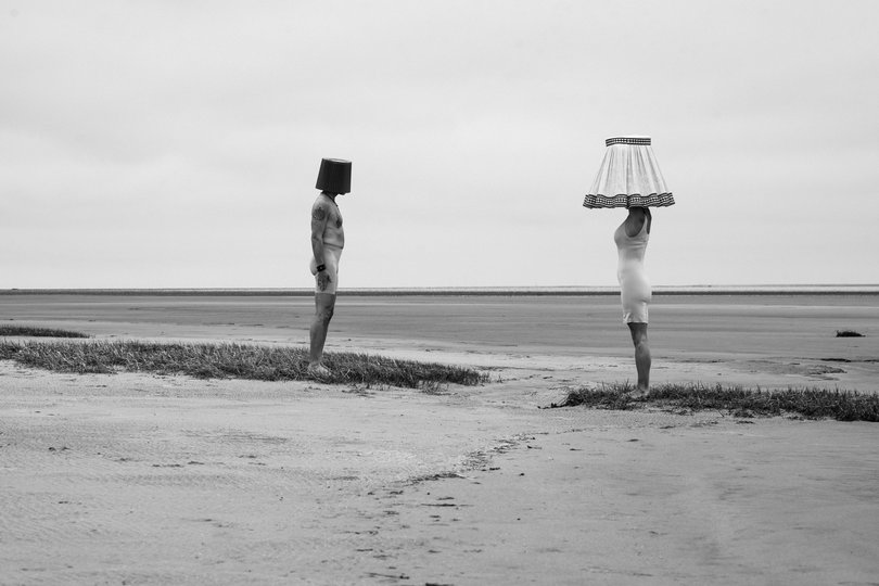 Ein Mann und eine Frau am Strand, sie tragen einen Lampenschirm über dem Kopf. Foto: Carola Bührmann