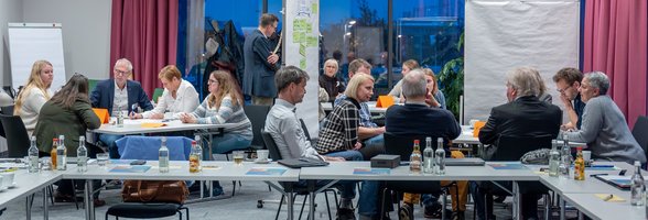 Menschen sitzen an drei Tischen zusammen und sprechen miteinander. Foto: plan zwei