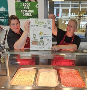 Petra Frerichs und Wenke Ommen zeigen das Plakat zur RegioWoche vor ihrer Essensausgabe Ein Speisenangebot in der Regiowoche 2025