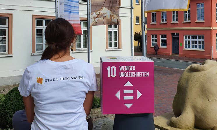 Frau mit dem Rücken zur Kamera und dem Logo der Stadt Oldenburg auf dem Rücken, unter dem Arm ein Pappwürfel mit dem SDG 10, im Hintergrund wehende Fahnen unter anderem von der Charta der Vielfalt. Foto: Stadt Oldenburg