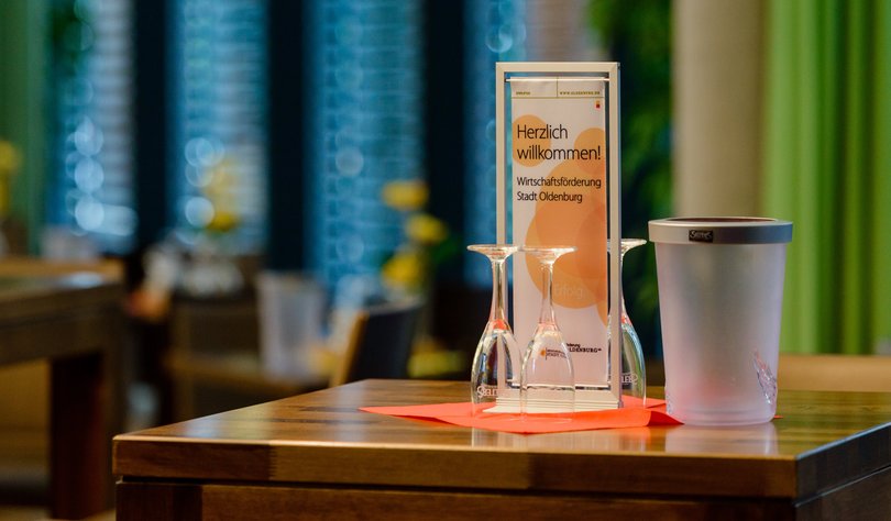 Tischdisplay mit Aufschrift „Herzlich willkommen! Wirtschaftsförderung Stadt Oldenburg“ auf einem Stehtisch in einem Veranstaltungssaal. Foto: Thorsten Helmerichs