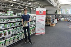 Timoté Caillarec bei Haushalt International. Foto: Stadt Oldenburg Timoté Caillarec steht vor einem Regal mit Produkten, er hält einen Karton. Foto: Stadt Oldenburg