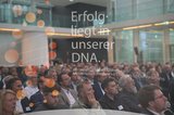Gäste eines Kontaktpunkts lauschen dem Keynote-Speaker. Im Vordergrund spiegelt sich ein Display der Wirtschaftsförderung mit der Aufschrift: „Erfolg liegt in unserer DNA“. Foto: Mohssen Assanimoghaddam