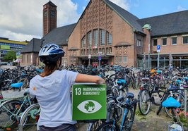 Frau mit Fahrradhelm mit dem Rücken zur Kamera und dem Logo der Stadt Oldenburg auf dem Rücken, unter dem Arm ein Pappwürfel mit dem SDG 13, im Hintergrund der Oldenburger Hauptbahnhof und dort geparkte Fahrräder Foto: Stadt Oldenburg