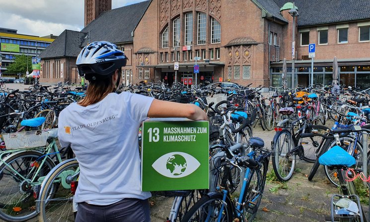 Frau mit Fahrradhelm mit dem Rücken zur Kamera und dem Logo der Stadt Oldenburg auf dem Rücken, unter dem Arm ein Pappwürfel mit dem SDG 13, im Hintergrund der Oldenburger Hauptbahnhof und dort geparkte Fahrräder Foto: Stadt Oldenburg