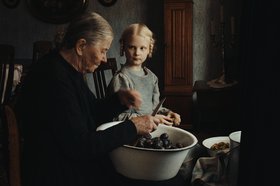Eine großmütterliche Frau sitzt auf einem Stuhl, eine große Schüssel mit Pflaumen auf ihrem Schoß, die sie schneidet. Daneben ein Mädchen mit blonden Zöpfen, das sie aufmerksam anschaut. Foto: Neue Visionen Filmverleih GmbH
