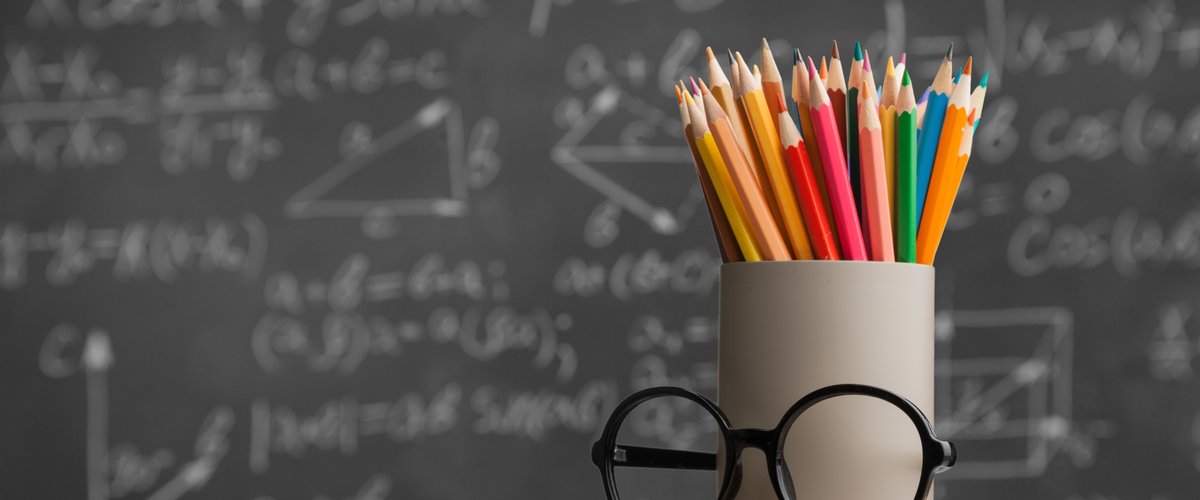 Brille und ein Köcher mit Stiften vor einer Tafel. Foto: BillionPhotos/AdobeStock