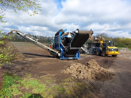 Die Säcke werden zunächst mit dem Bagger in den Schredder befördert, anschließend wird das Material durch die blaue Siebtrommel getrennt. Am Ende entstehen Haufen mit Sackmaterial aus Jute (vorne) beziehungsweise Sand (links). Foto: Stadt Oldenburg