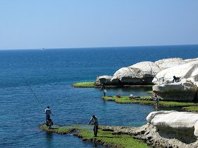 Blaues Meer, blauer Himmel und weiße Felsen. Am Ufer stehen zwei Menschen und angeln. Foto: Matte Asher Regional Council