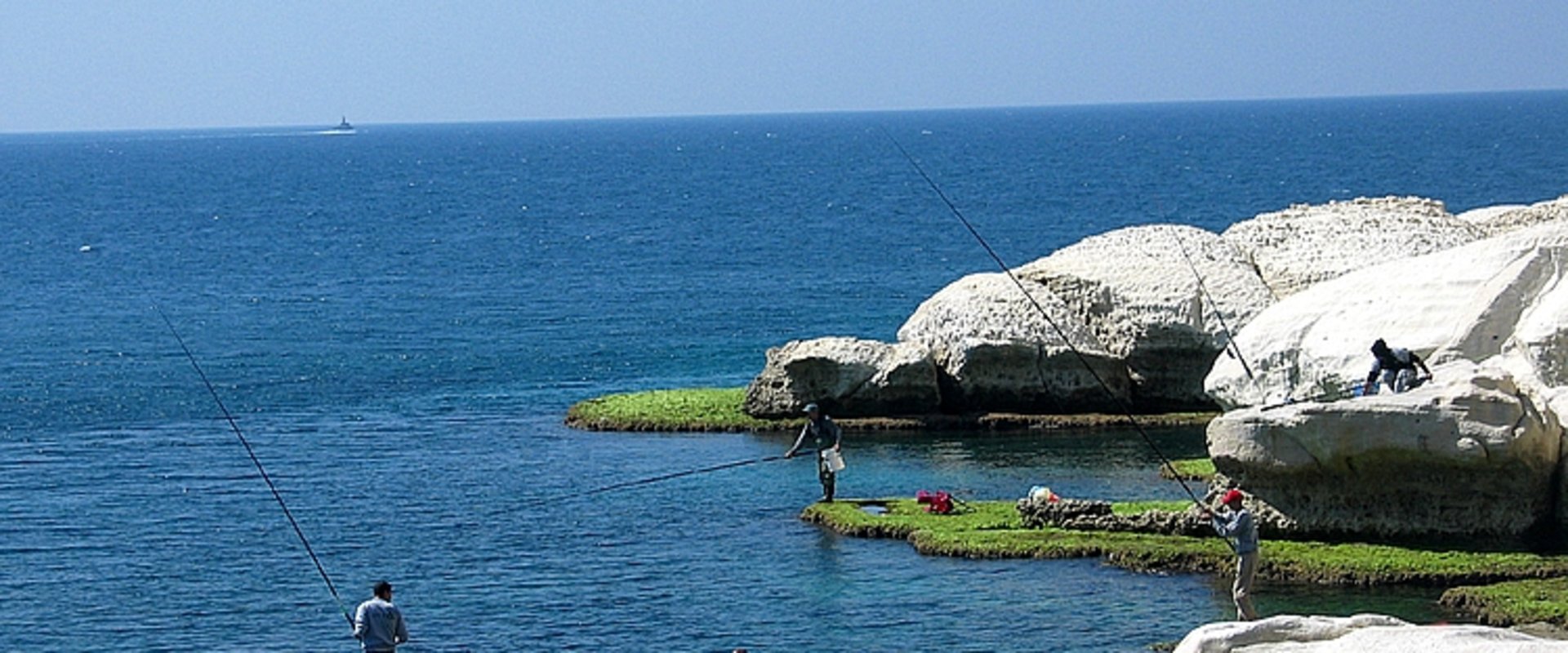 Blaues Meer, blauer Himmel und weiße Felsen. Am Ufer stehen zwei Menschen und angeln. Foto: Matte Asher Regional Council
