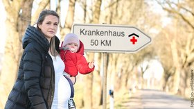 Frau mit Kind auf dem Arm. Im Hintergrund ein Schild mit der Aufschrift „Krankenhaus 50 km“. Foto: Foto- und Bilderwerk