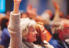 Menschen auf einer Versammlung, eine Seniorin meldet sich. Foto: GoshaMovies/AdobeStock