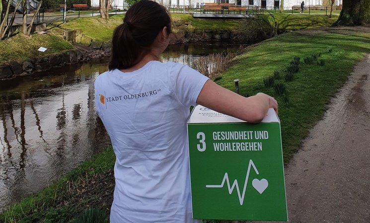 Frau mit dem Rücken zur Kamera und dem Logo der Stadt Oldenburg auf dem Rücken, unter dem Arm ein Pappwürfel mit dem SDG 3, im Hintergrund Gebäude des Pius Hospitals am Wallgraben. Foto: Stadt Oldenburg