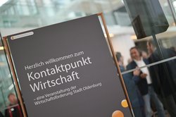 Im Vordergrund: ein Banner der Wirtschaftsförderung mit dem Aufdruck „Herzlich willkommen zum Kontaktpunkt Wirtschaft – eine Veranstaltung der Wirtschaftsförderung Stadt Oldenburg.“ Im Hintergrund: Gäste des Kontaktpunkts vernetzen sich. Foto: Mohssen Assanimoghaddam