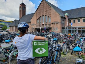 Frau mit Fahrradhelm mit dem Rücken zur Kamera und dem Logo der Stadt Oldenburg auf dem Rücken, unter dem Arm ein Pappwürfel mit dem SDG 13 (Maßnahmen zum Klimaschutz), im Hintergrund der Oldenburger Hauptbahnhof und dort geparkte Fahrräder Foto: Stadt Oldenburg