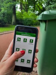 Hand mit Handy, auf dem die AWB-App geöffnet ist. Im Hintergrund eine grüne Tonne. Foto: Stadt Oldenburg