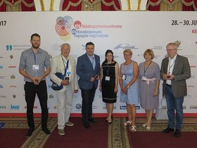 Steffen Trawinski, Siegfried Dose, Dragos Pancescu, eine Mitarbeiterin der Stadtverwaltung Woronesch, Marianne Vieler-Bargfeldt, Emilia Winterholler und Helmut Hinrichs stehen vor einer Wand mit der Aufschrift „14. Deutsch-Russische Städtepartnerkonferenz“. Foto: Stadt Oldenburg