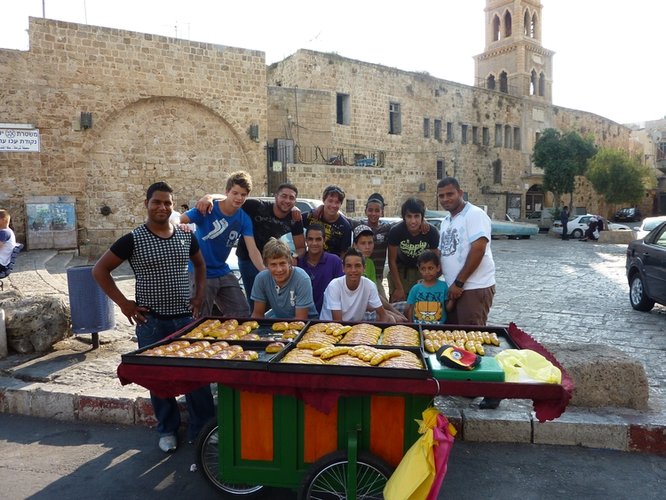 Eine Gruppe Jugendliche steht an einem Verkaufsstand mit Gebäck, im Hintergrund ist ein Stadtmauer aus hellem Stein zu sehen. Foto: Stadt Oldenburg