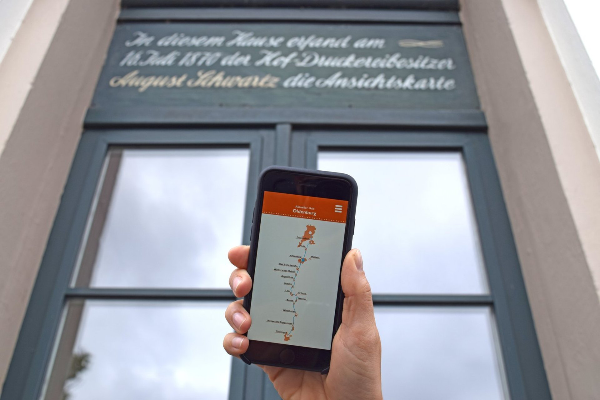 Smartphone mit der Wunderline-App vor dem Haus des Schlossplatzes 21/22, in dem Hof-Druckereibesitzer August Schwartz die Ansichtskarte erfand. Foto: Stadt Oldenburgr August Schwartz 1870 die Ansichtskarte erfand. Foto: Stadt Oldenburg 