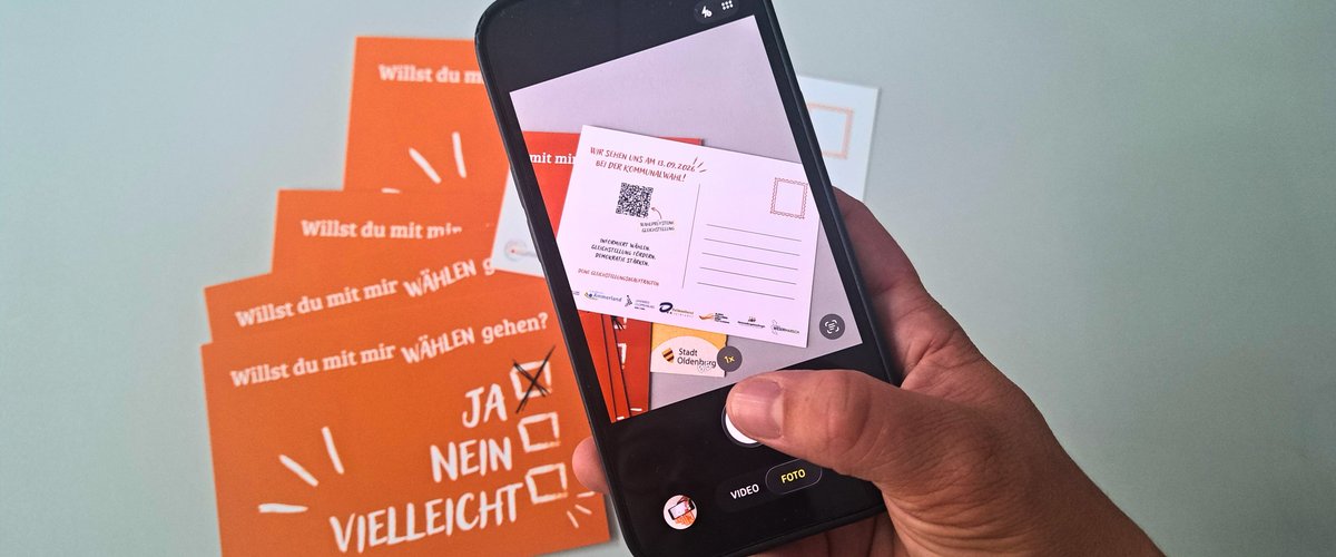 Eine Hand hält ein Smartphone und fotografiert die orangenen Postkarten mit der Aufschrift „Willst du mit mir wählen gehen? – Ja. Nein. Vielleicht.“. Foto: Stadt Oldenburg