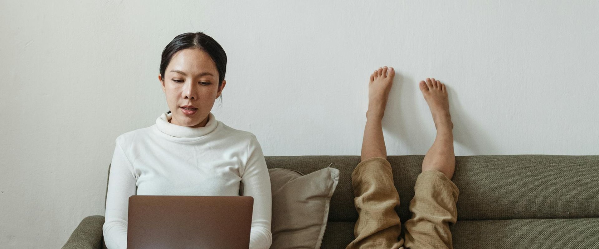Eine Frau arbeitet auf einem Sofa an einem Laptop, daneben liegt ein Kind mit dem Rücken auf der Sitzfläche und streckt die Beine an der Rückenlehne hoch. Foto: pexels/Ketut Subiyanto