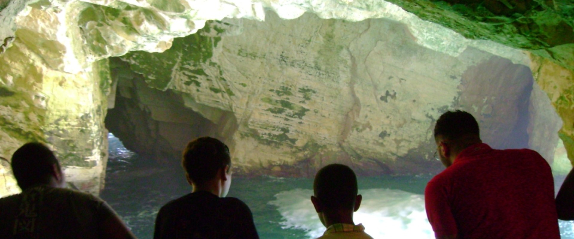 Vier Jugendliche von hinten aufgenommen, im Hintergrund eine Grotte aus weißem Fels, unten ist türkisfarbenes Wasser zu sehen. Foto: Edna Taff
