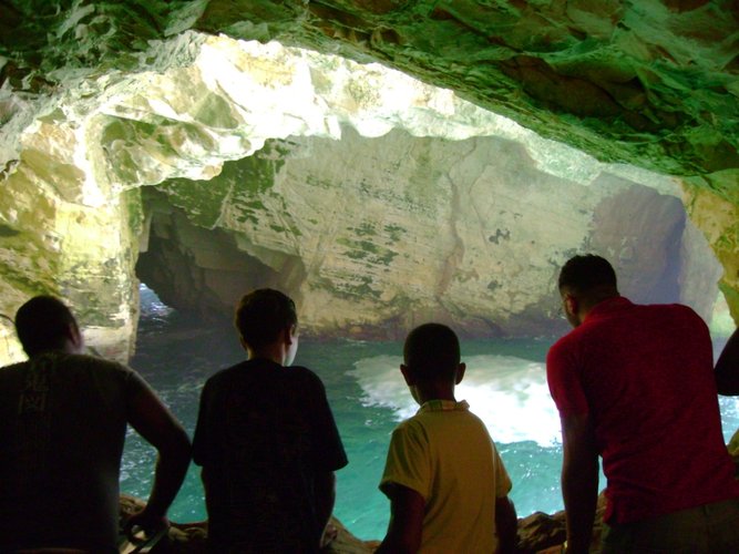 Vier Jugendliche von hinten aufgenommen, im Hintergrund eine Grotte aus weißem Fels, unten ist türkisfarbenes Wasser zu sehen. Foto: Edna Taff